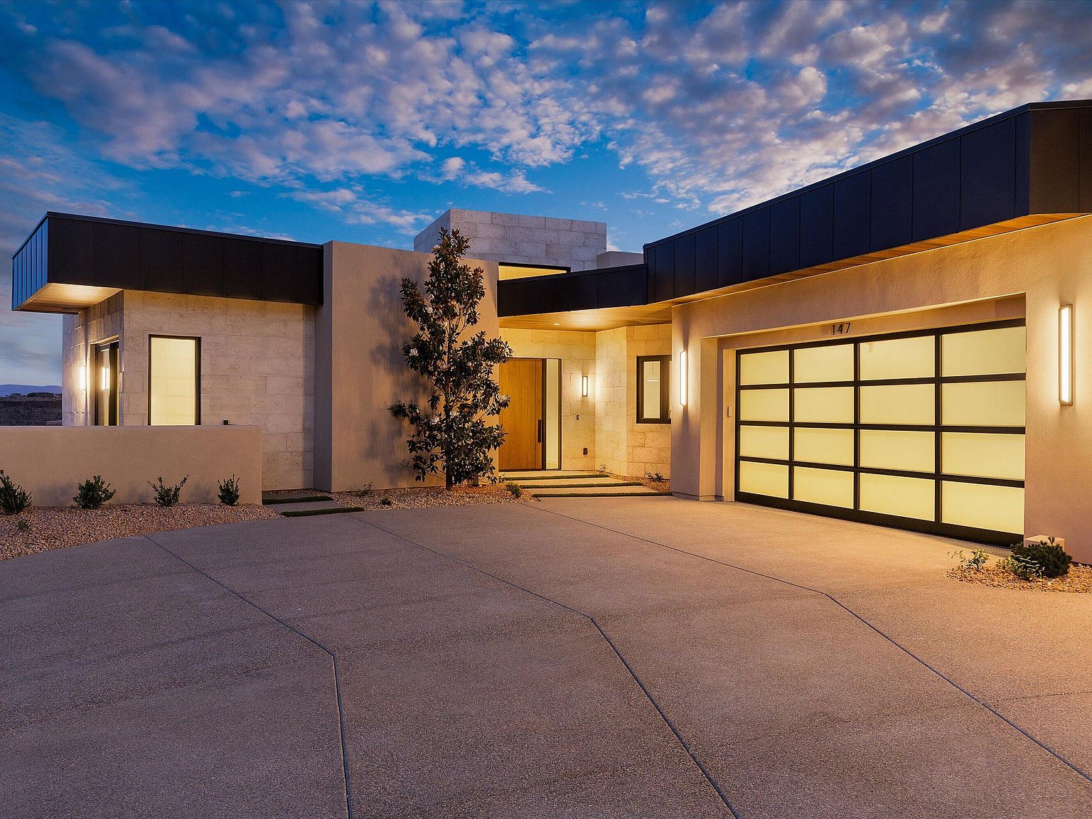 Twilight front elevation with mature trees framing the limestone entry, illuminated glass garage, and the address number 147 above the door