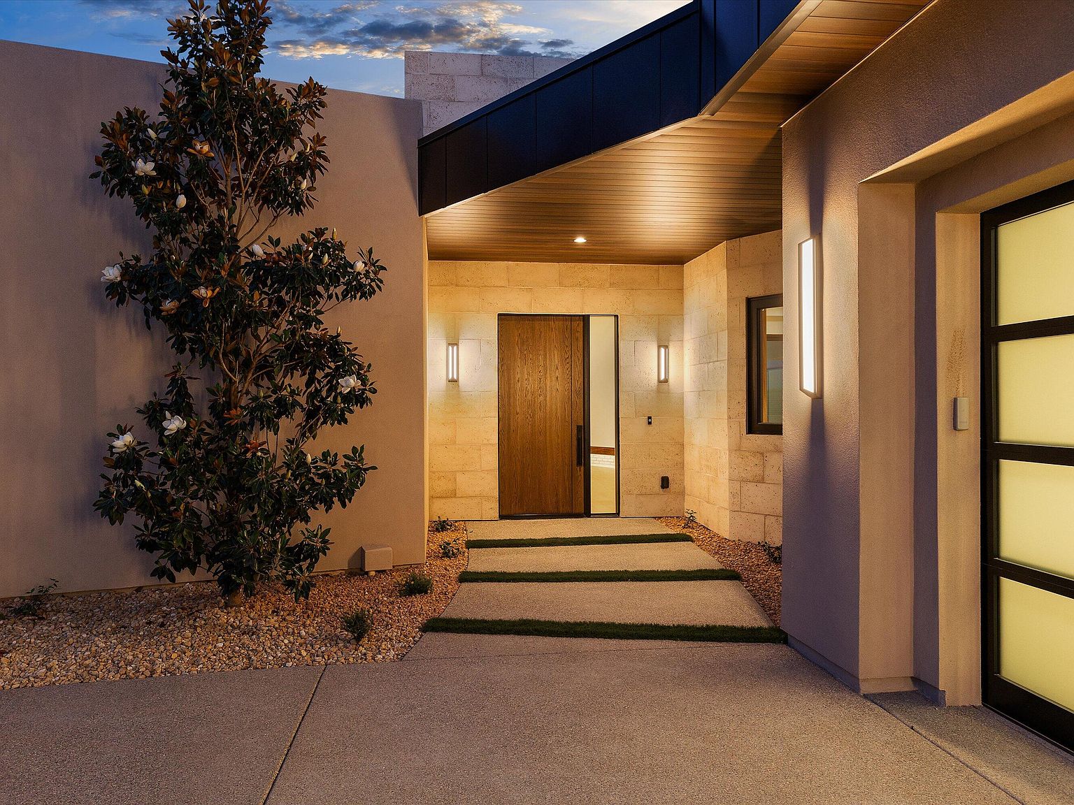 Twilight entry courtyard showing the custom wood front door flanked by sconces, framed by a mature magnolia tree and turfed concrete walkway