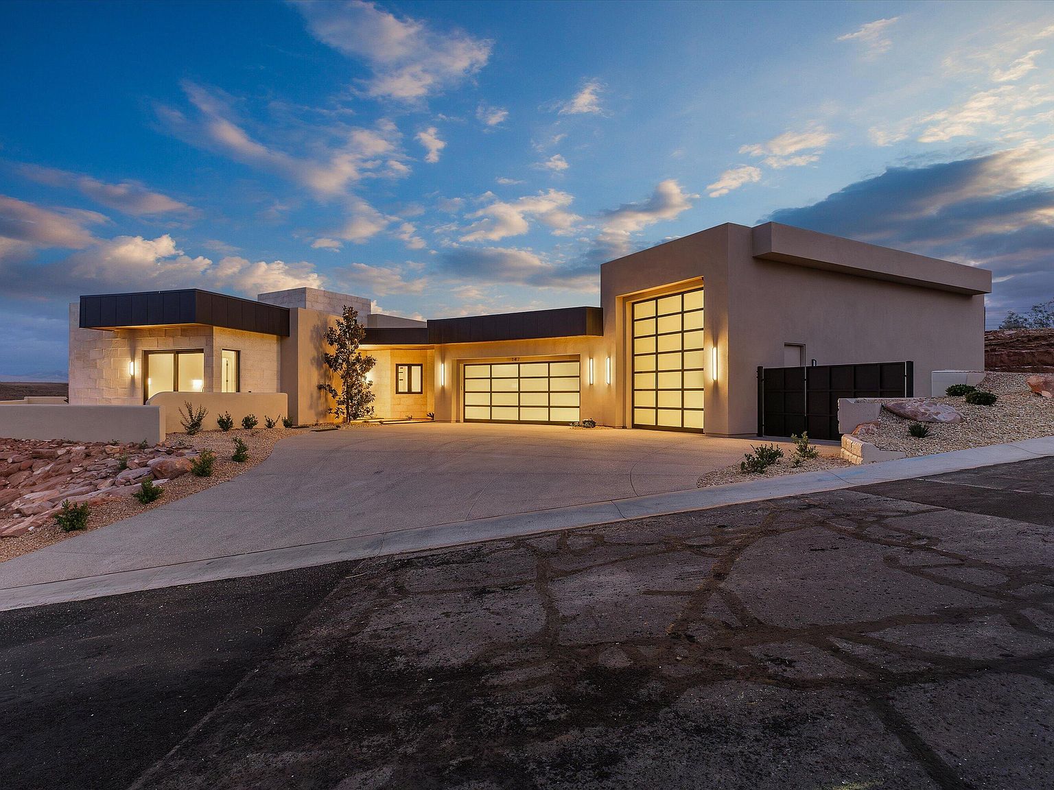 Twilight front elevation of 147 N Westgate Hills Drive, a single-level modern desert residence with custom limestone, dark fascia, and a glass-paneled three-car garage glowing against a southern Utah dusk sky